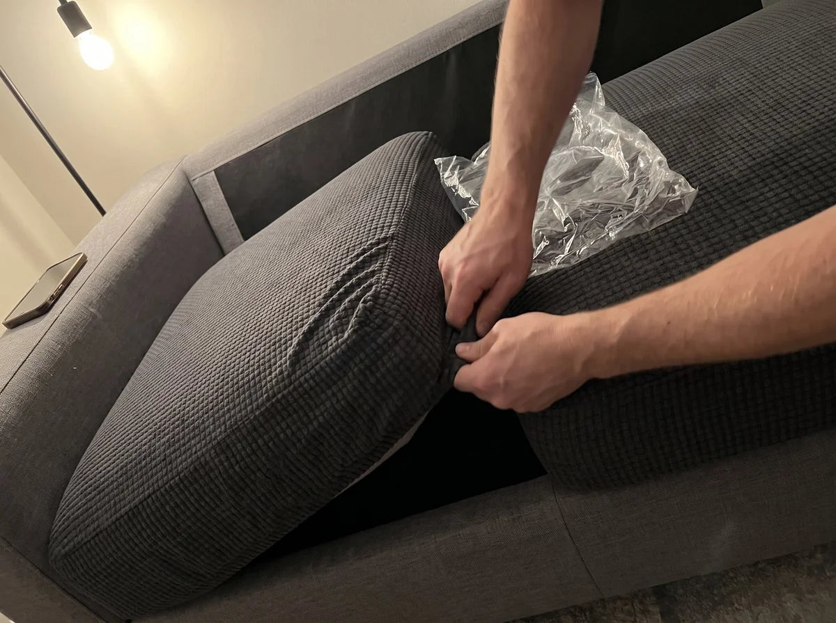 Hands tucking the elastic hem of a dark grey Avelo Casa seat cover under a sofa cushion